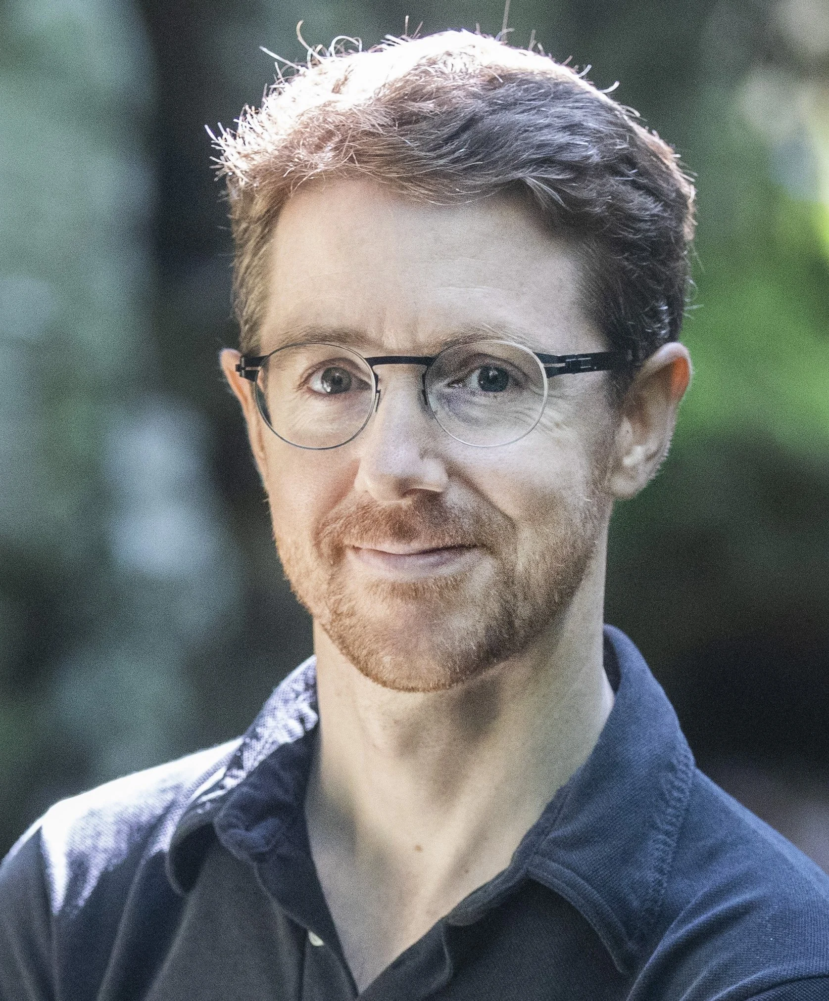 A man with glasses and a beard outdoors, smiling with blurred greenery in the background.