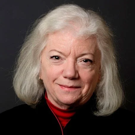 An elderly woman with gray hair smiling, wearing a black top and red turtleneck, against a dark background.