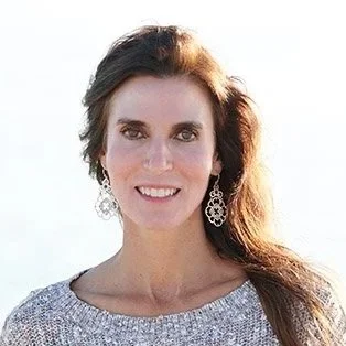 A woman with long brown hair smiling, wearing earrings and a light-colored top, outdoors with a bright sky background.