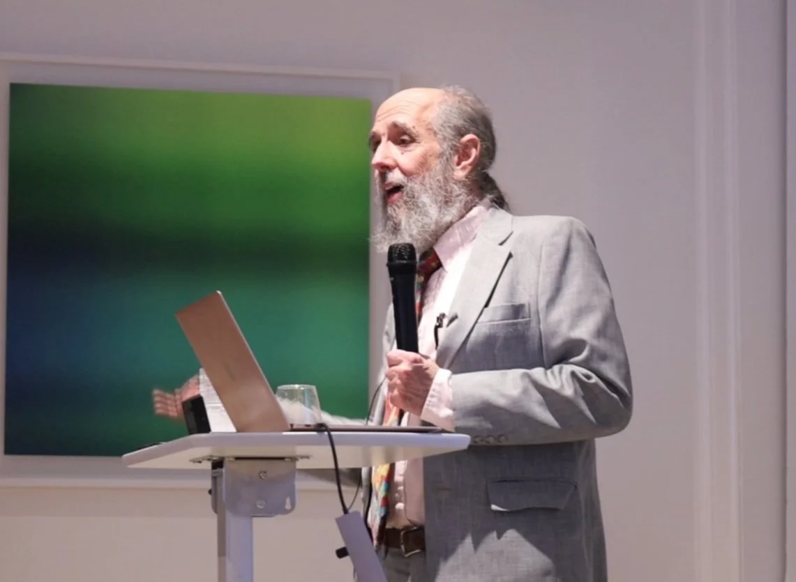 An older man with a gray beard and long hair in a ponytail, wearing a light gray suit, is giving a presentation. He is holding a microphone in his right hand and standing behind a stand with a laptop and some papers. There is a glass of water on the stand. The background shows a colorful abstract painting on a white wall.