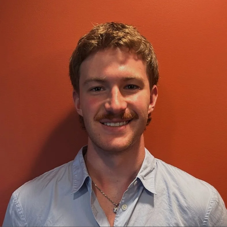 A young man with light brown hair, a mustache, wearing a light blue button-up shirt and a silver necklace, smiling against an orange wall.