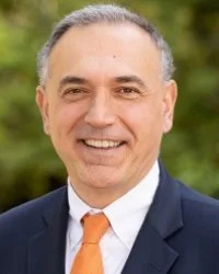 A man with short gray hair smiling in front of a green outdoor background, wearing a dark suit, white shirt, and orange tie.