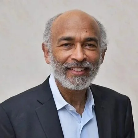 Headshot of a man with a gray beard and dark blazer, smiling warmly against a neutral background.