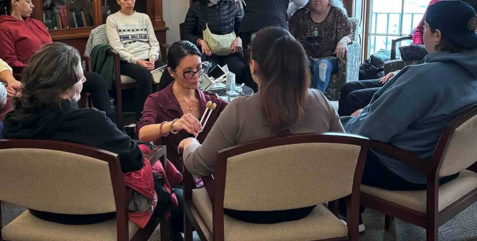 A group of women seated in a circle of chairs engaged in a therapy or support group session, with one woman offering comforting touch.