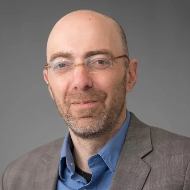 Portrait of a bald man wearing glasses, a blue shirt, and a gray blazer, against a gray background.
