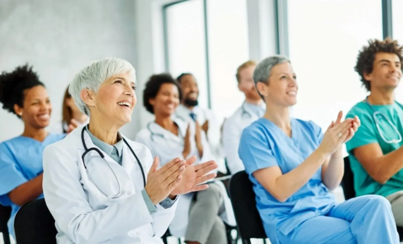 Group of healthcare professionals, including doctors and nurses, sitting and clapping in a brightly lit room during an event or presentation.
