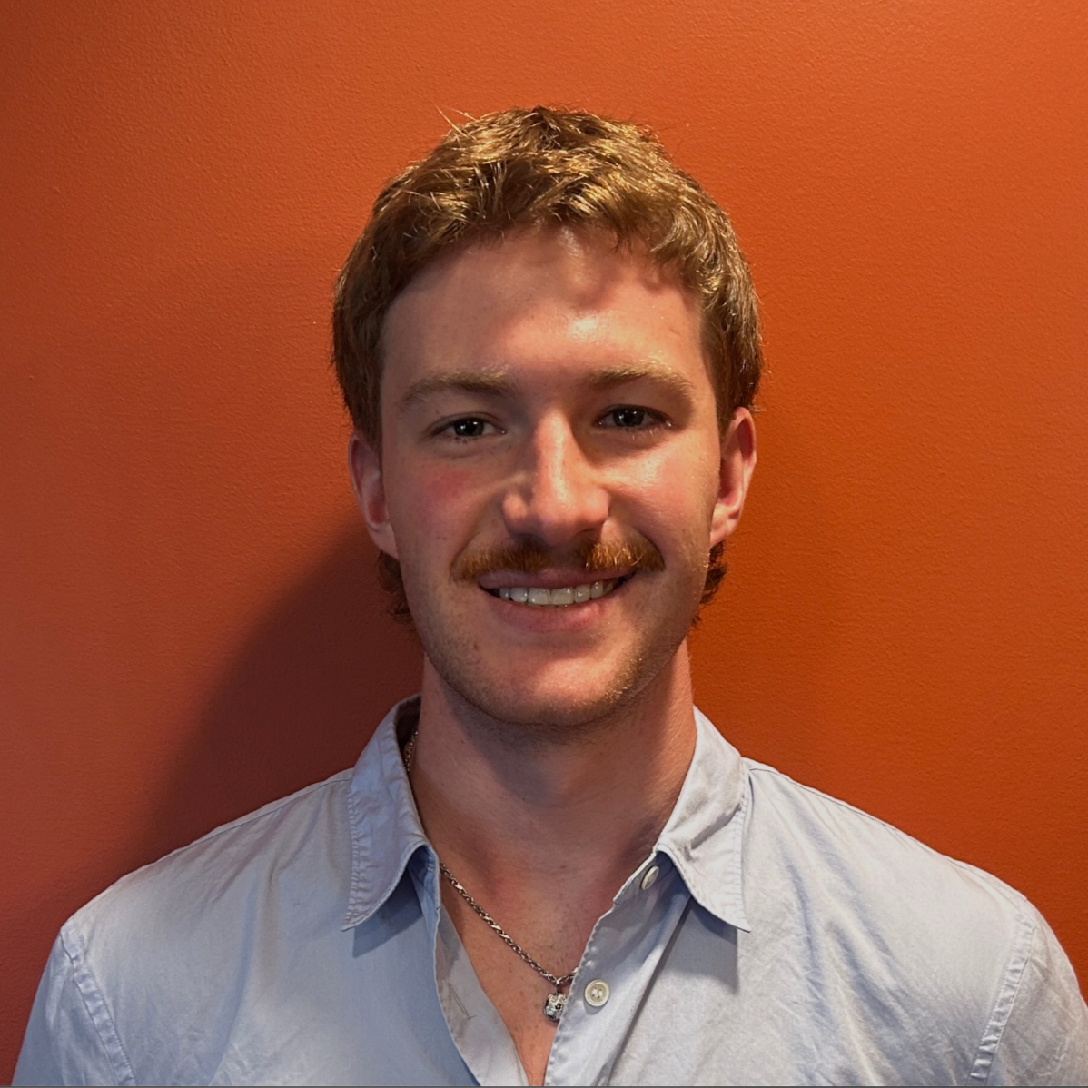 A young man with light brown hair, a mustache, wearing a light blue button-up shirt and a silver necklace, smiling against an orange wall.