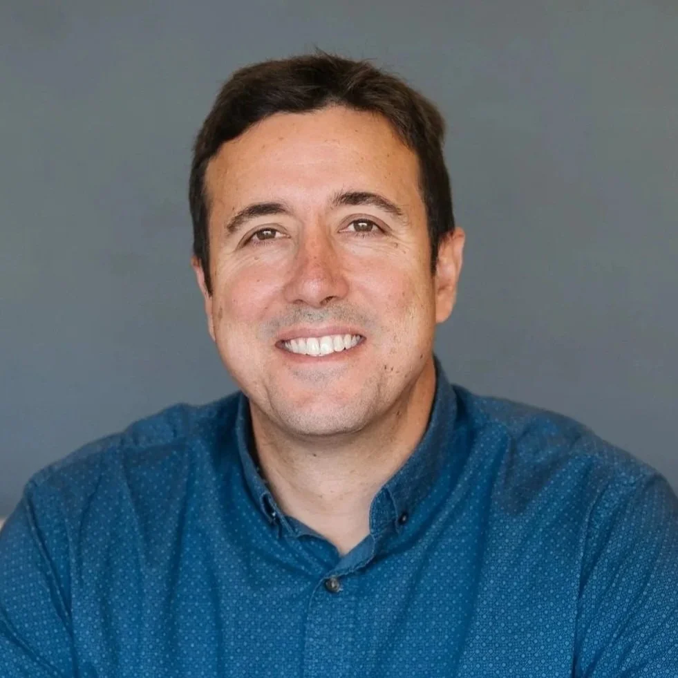 A smiling man with dark hair and light skin, wearing a blue button-up shirt, standing against a gray background.