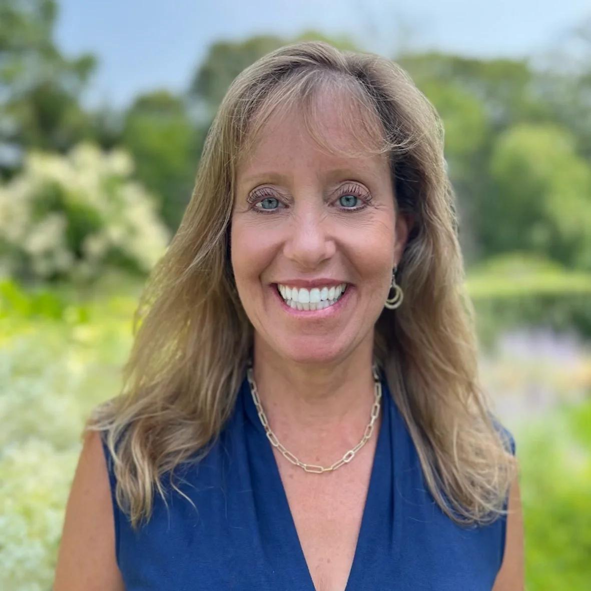 A smiling woman with light brown hair, blue eyes, wearing a blue sleeveless top, gold jewelry, outdoors with trees and a body of water in the background.