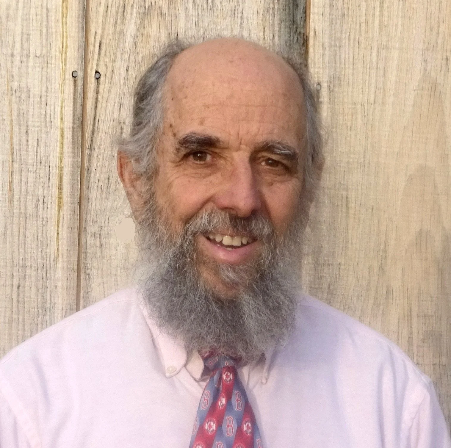 A smiling elderly man with a graying beard and mustache, wearing a light pink shirt and a colorful tie, standing in front of a wooden background.