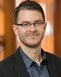 Headshot of a man with glasses and a dark blazer, smiling against a warm blurred background.