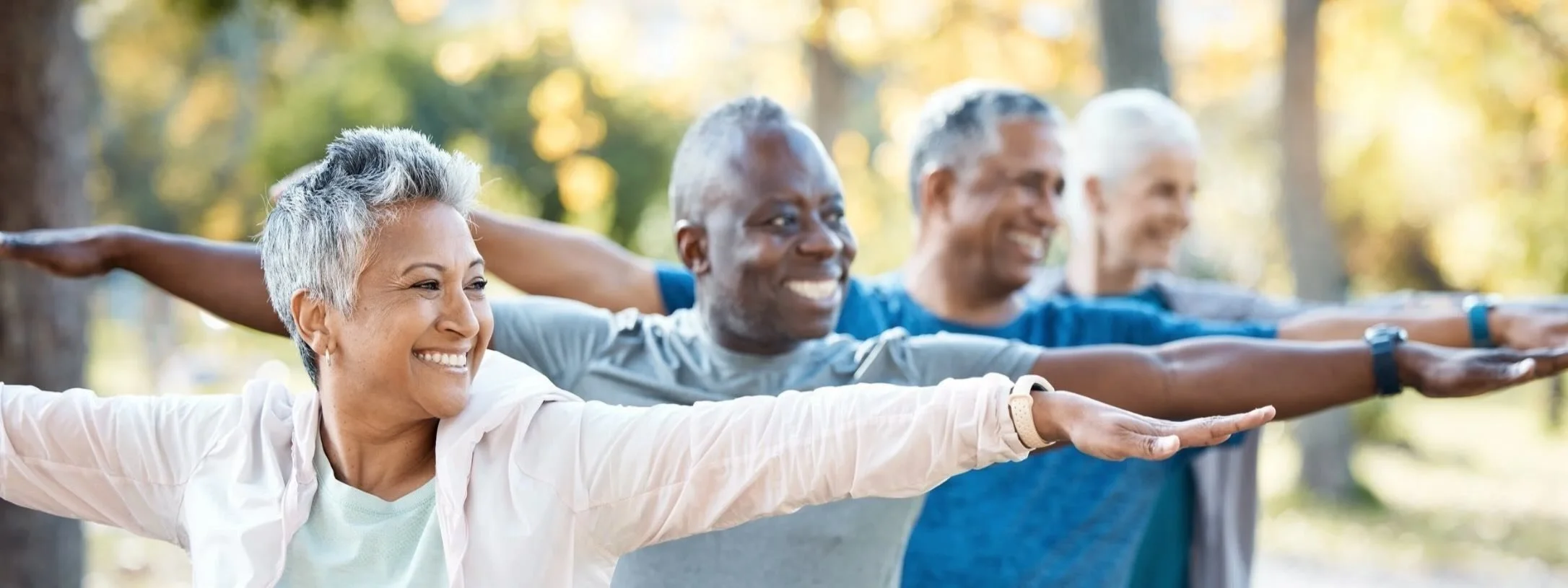 Group of four seniors practicing yoga outdoors in a park on a sunny day, arms extended.