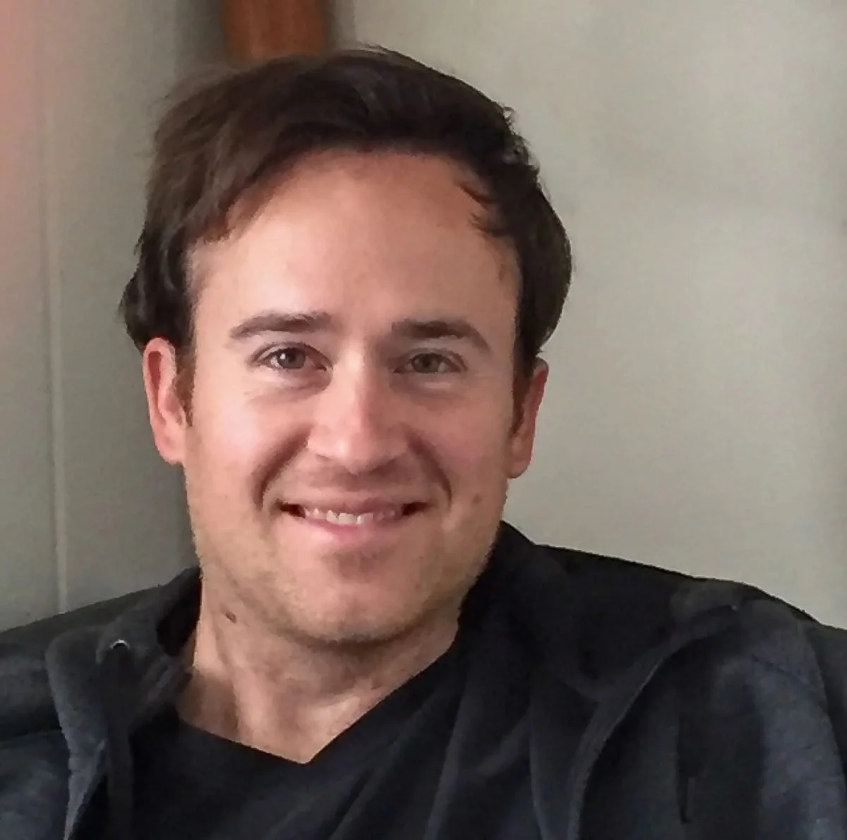A smiling man with dark hair, wearing a black jacket, sitting indoors near a wall with a wooden element in the background.