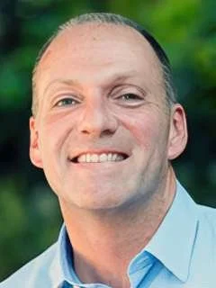 Portrait of a man with short hair, smiling, wearing a light blue shirt, outdoors with a green blurred background.