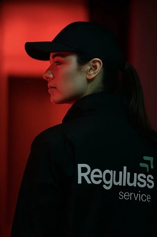 Frau in schwarzer Uniform mit schwarzer Kappe, Rücken und Profil sichtbar, steht vor einem roten Licht, auf der Jacke steht 'Reguluss service'.