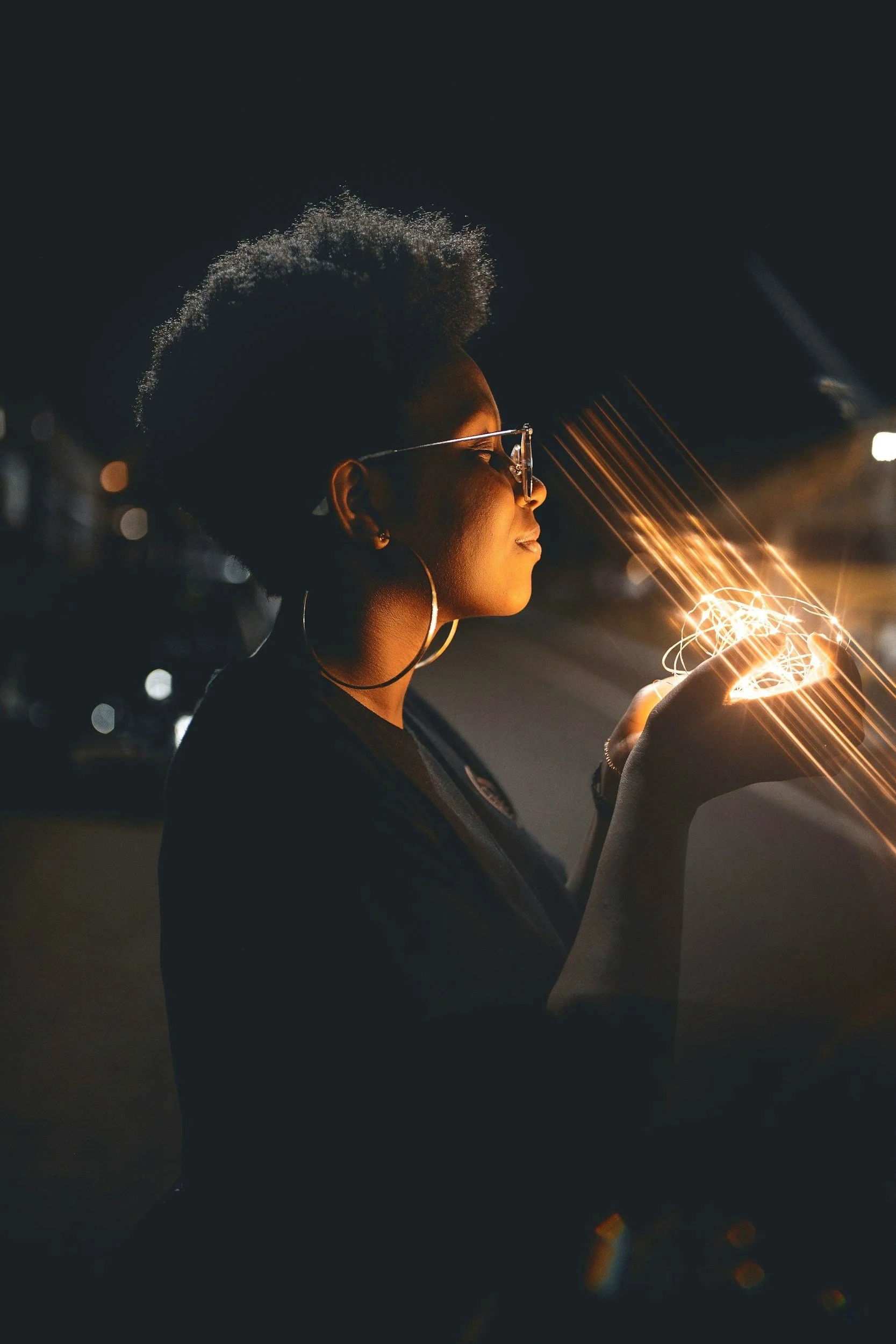 A black woman with short curly hair and large hoop earrings is illuminated by a glowing light from twinkle lights in her hands at night, wearing glasses - representing clear vision and strategic insight.
