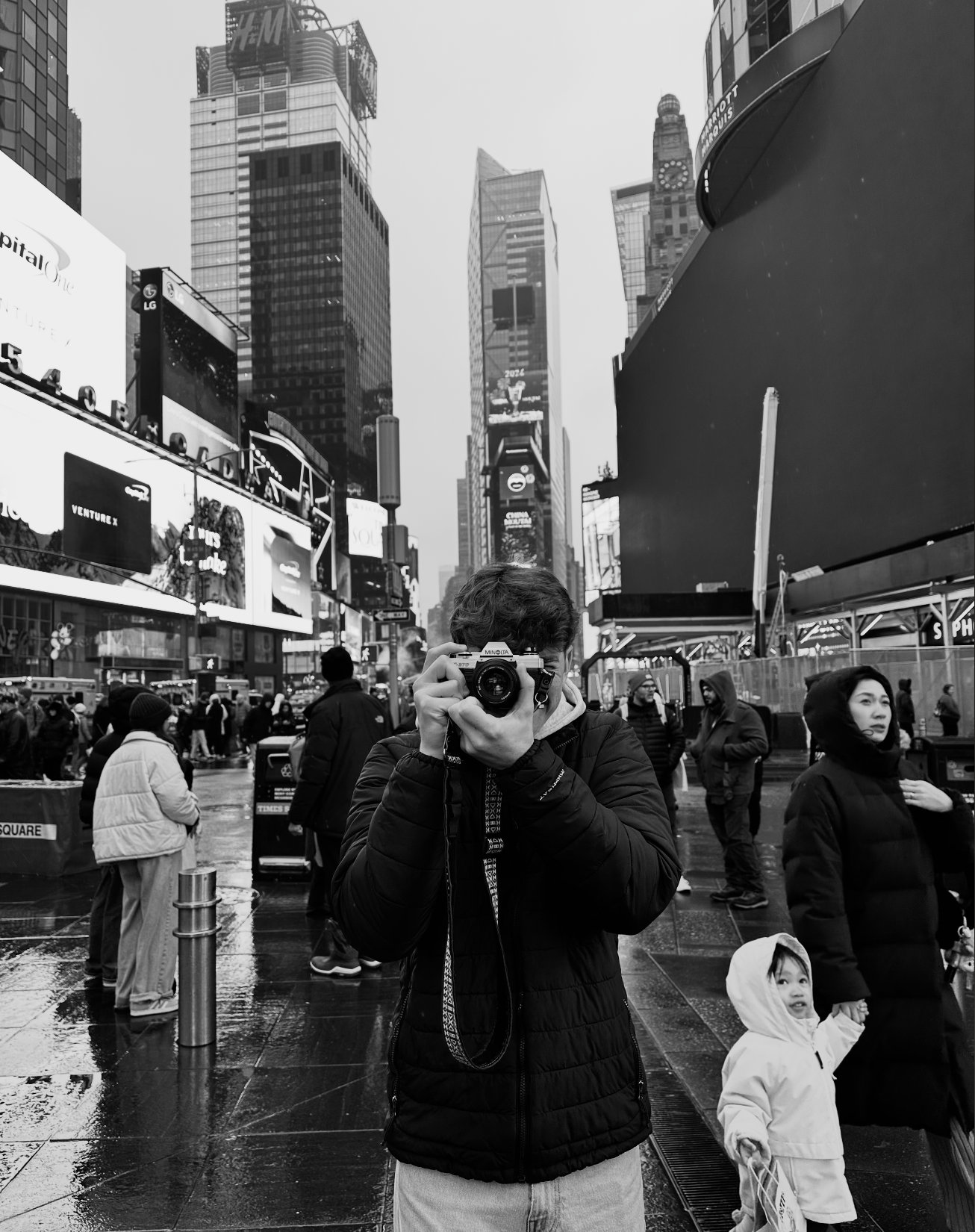 A person takes a photo with a camera in Times Square, New York City, on a rainy day, with tall buildings and digital billboards in the background. Other pedestrians and a child are visible around.