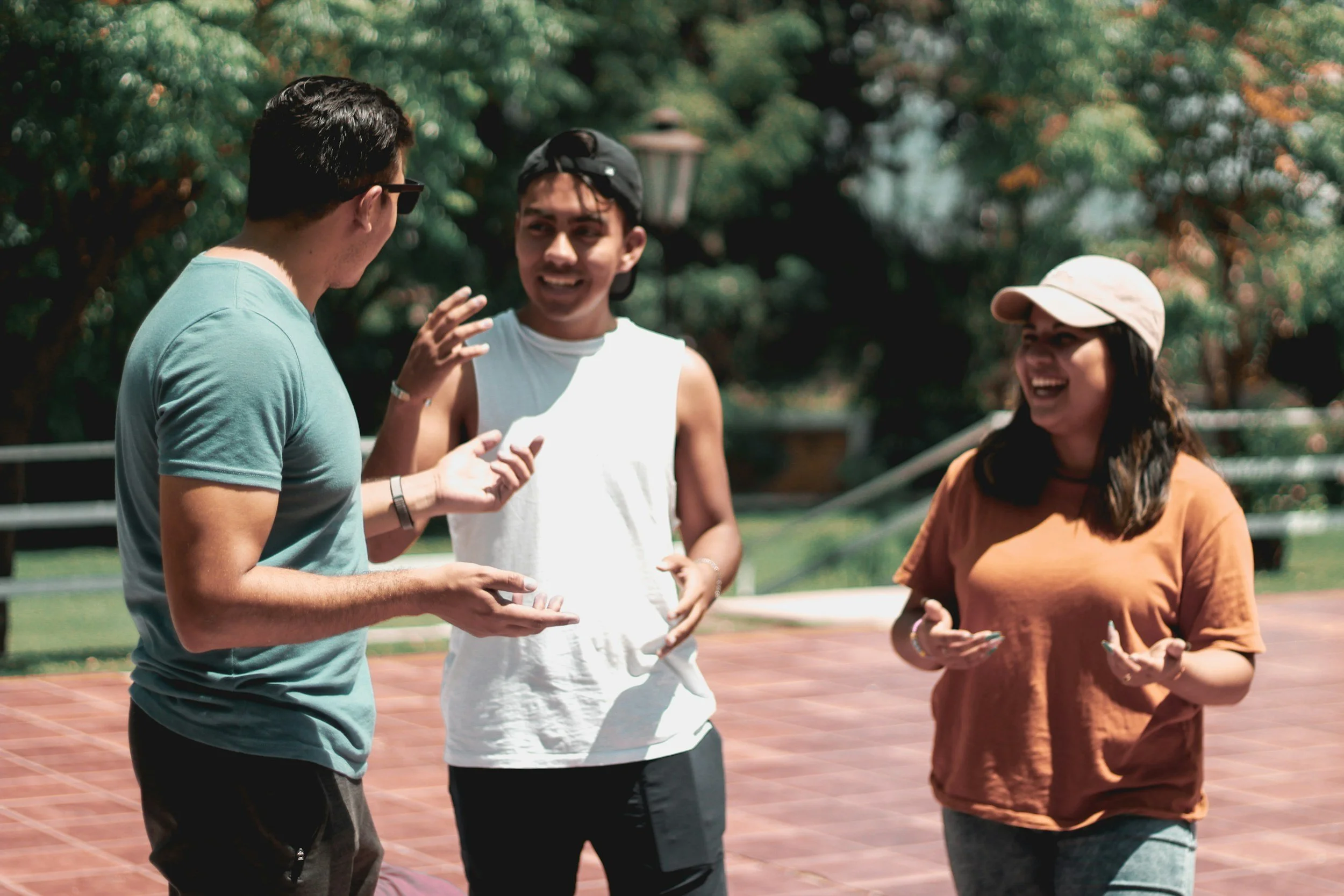 Three young people talking and smiling outdoors on a sunny day, standing on a tennis court surrounded by trees.