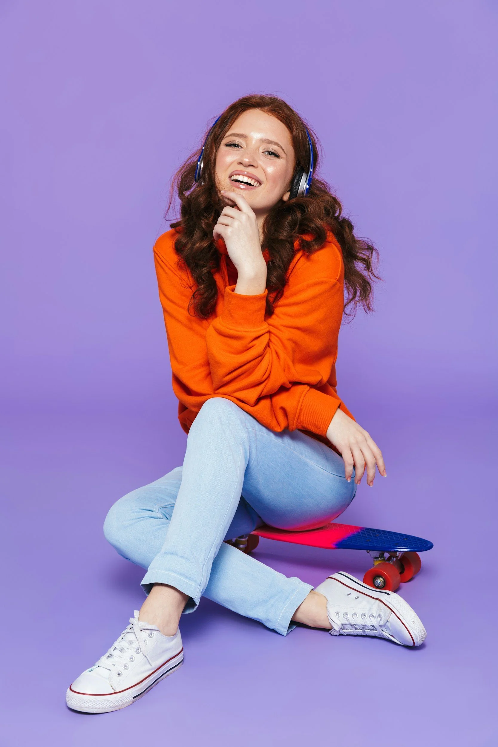 A young woman with curly red hair, wearing an orange sweater and light blue jeans, sitting on the floor with one leg crossed over the other, smiling while wearing headphones. A skateboard is beneath her. The background is purple.