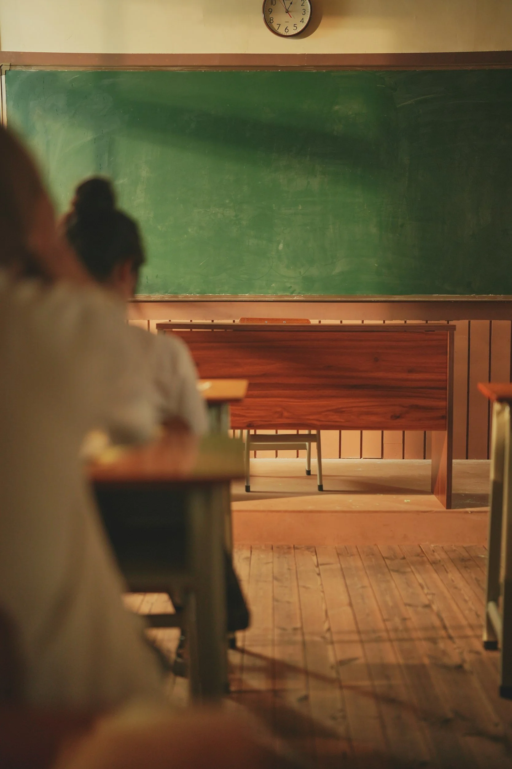 Scene of a classroom with a green chalkboard, wooden desks, and a clock on the wall showing 12:14.