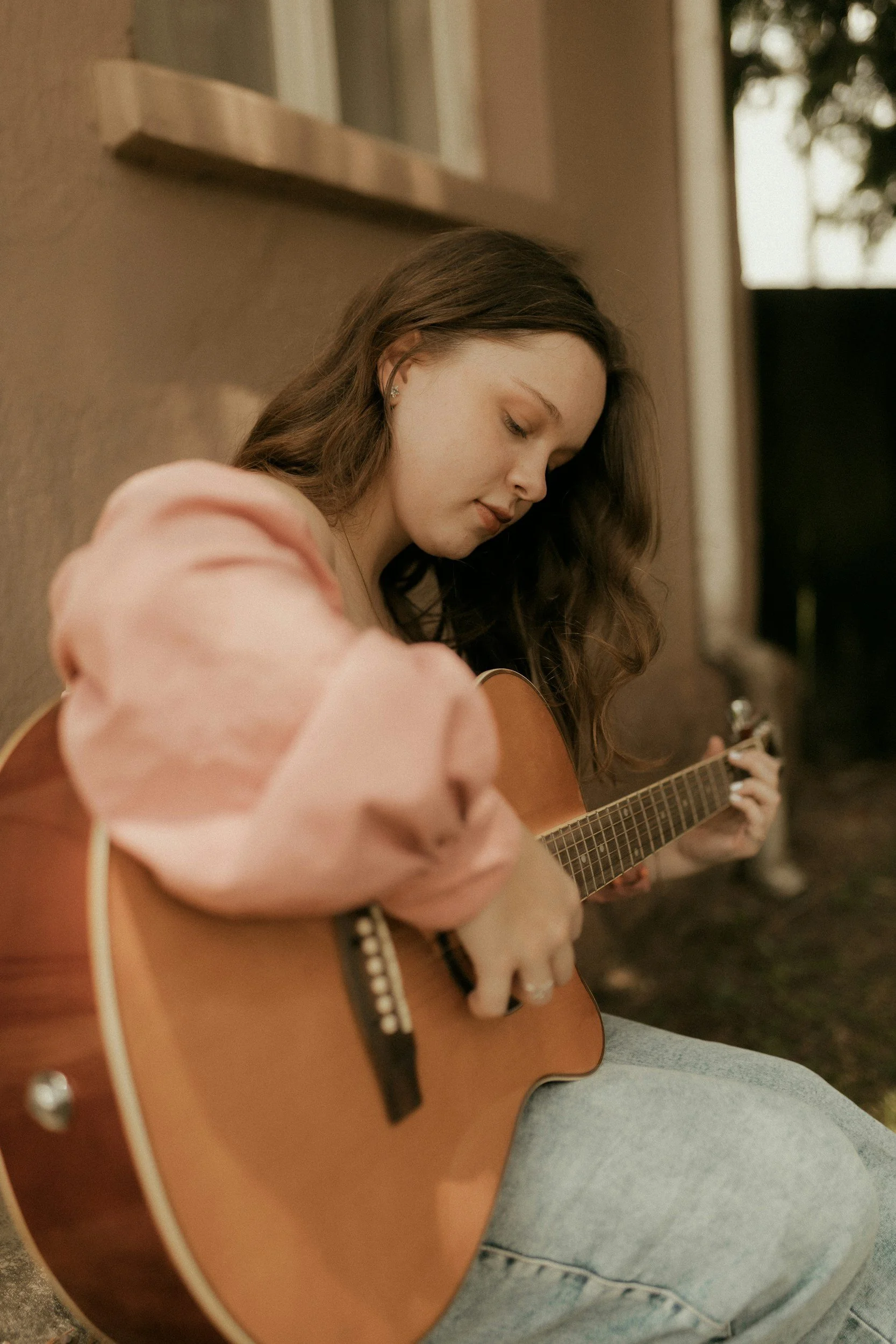 A young woman with long, wavy brown hair playing an acoustic guitar outdoors near a beige building with a window, wearing a pink top and gray jeans.