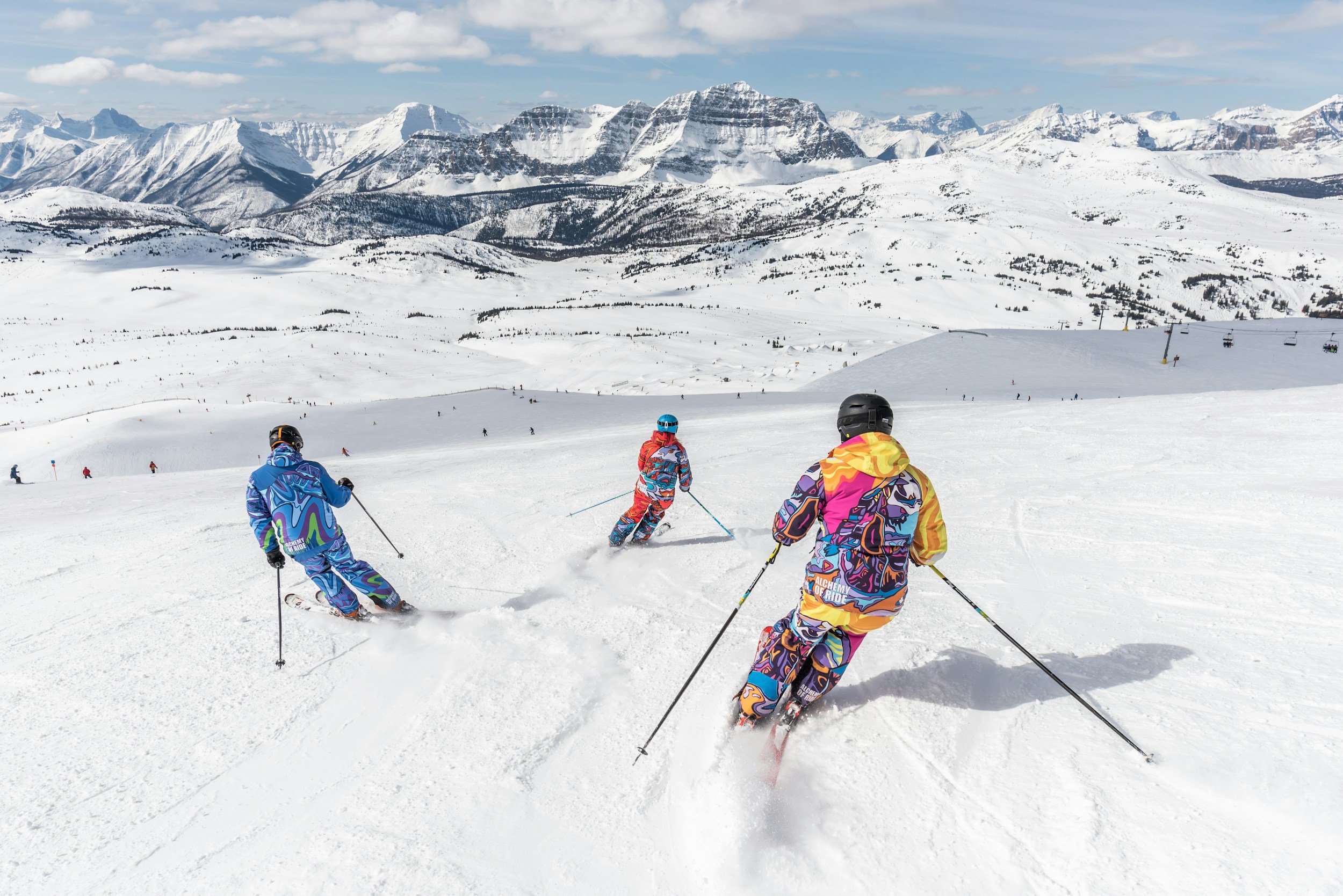 Three skiers dressed in colorful winter gear skiing down a snow-covered mountain slope with a vast snowy mountain range in the background under a partly cloudy sky.