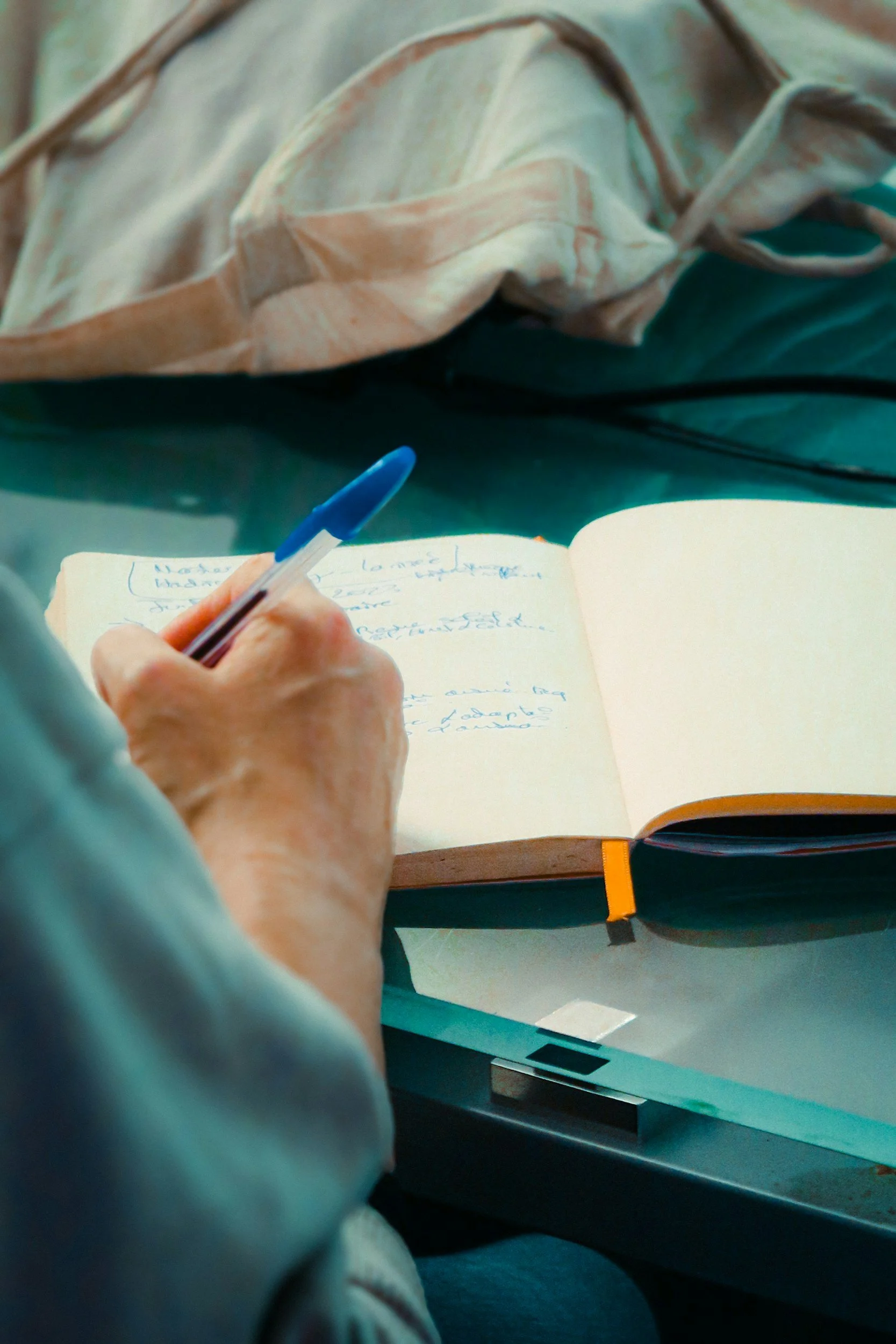 A person writing notes in a notebook with a blue pen, with a fabric item and a closed book on a desk.