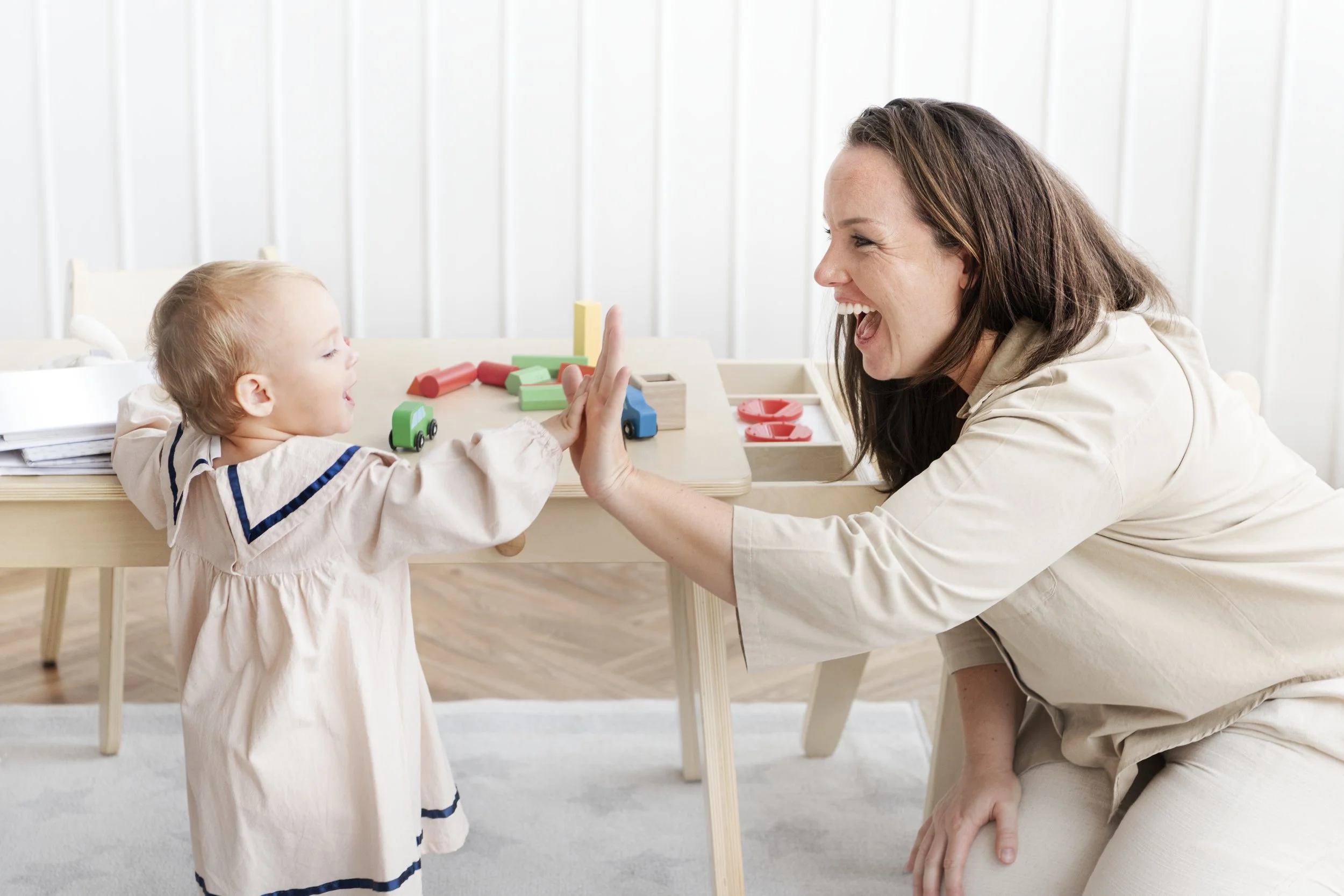 A young girl giving a high five to a woman, both smiling, at a table with colorful toy cars and blocks in a bright, minimalistic room.