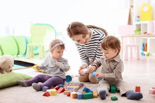 A woman playing with two young children on a carpeted floor surrounded by colorful toys.