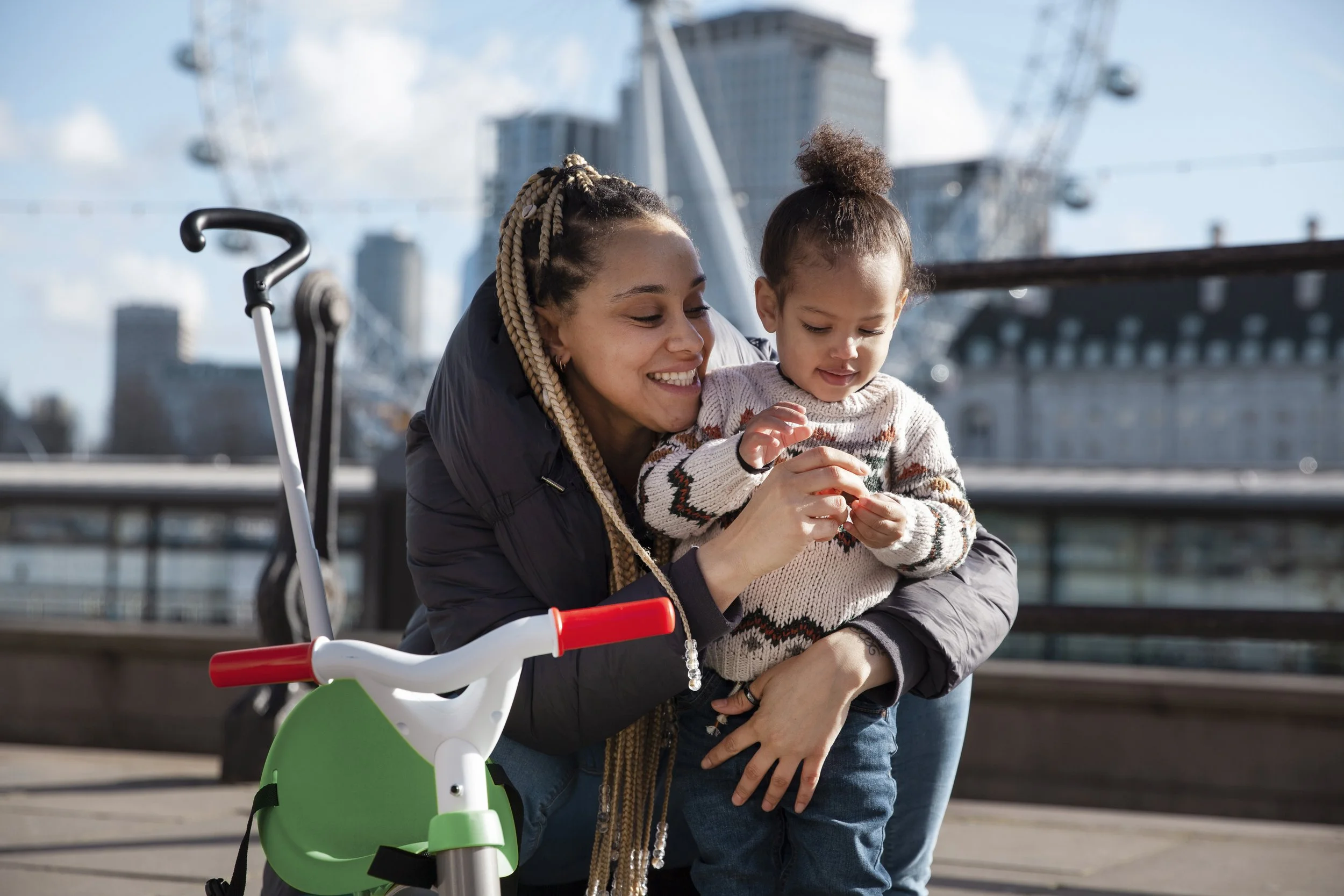 A woman with braided hair smiling and holding a young girl with curly hair in her arms, outdoors near a city waterfront with a Ferris wheel and skyscrapers in the background.