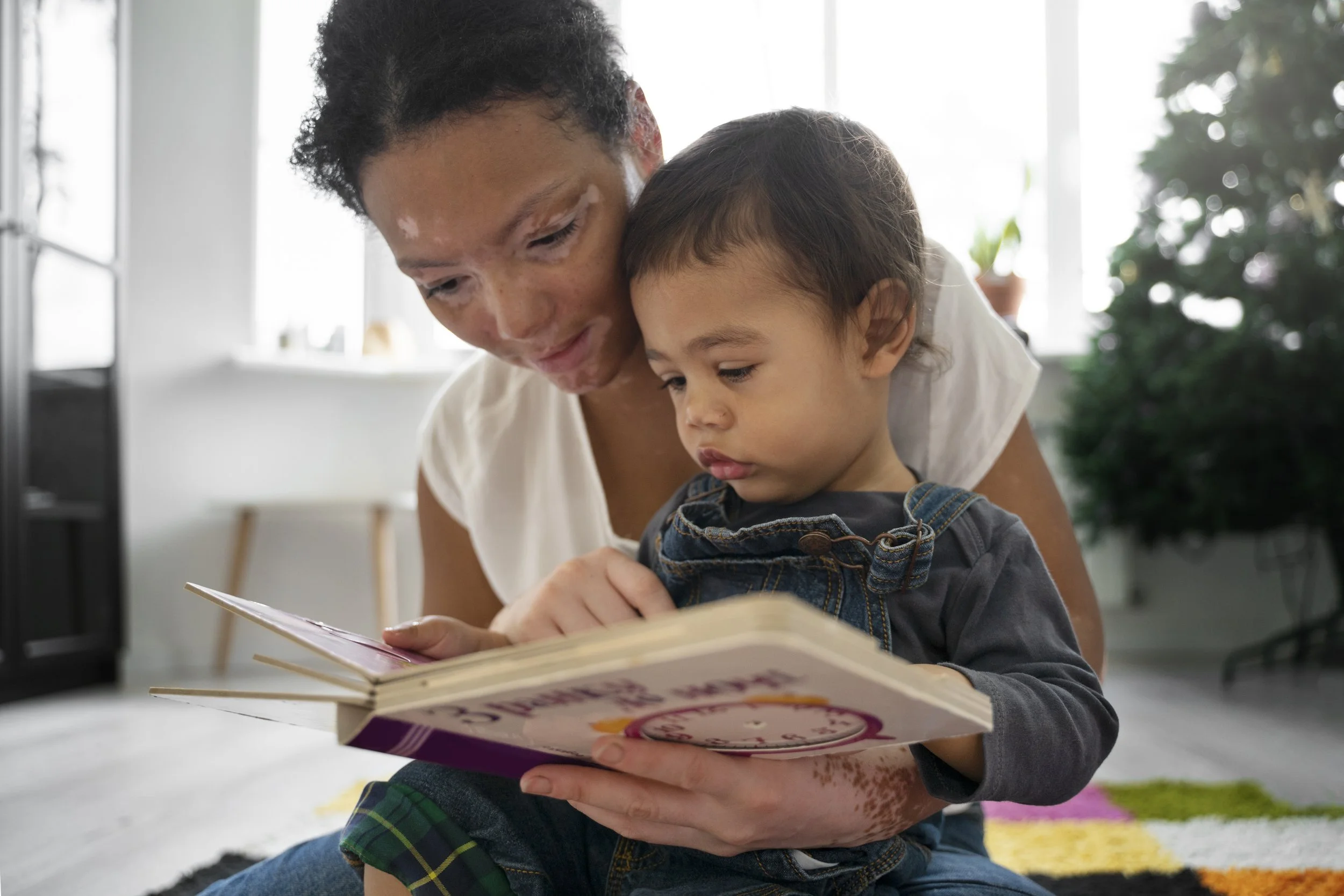 A woman with short curly hair and a young boy with short dark hair are sitting at a table, looking at a book together. The woman is smiling, and the boy is focused on the book. The background shows bright windows, and there is a plant to the right.