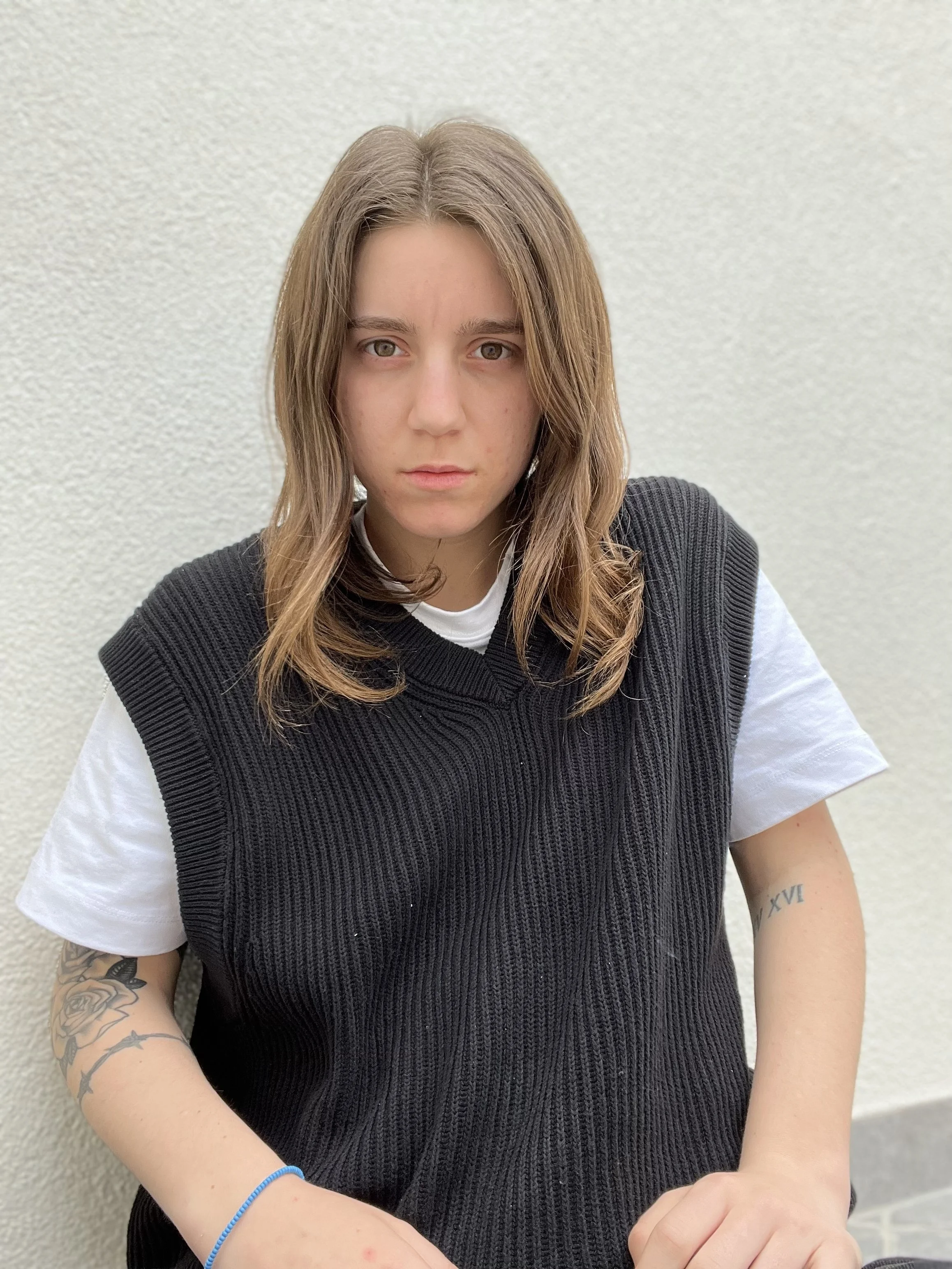 Ragazza con capelli castani, abbigliamento casual e tatuaggi, seduta contro un muro bianco.