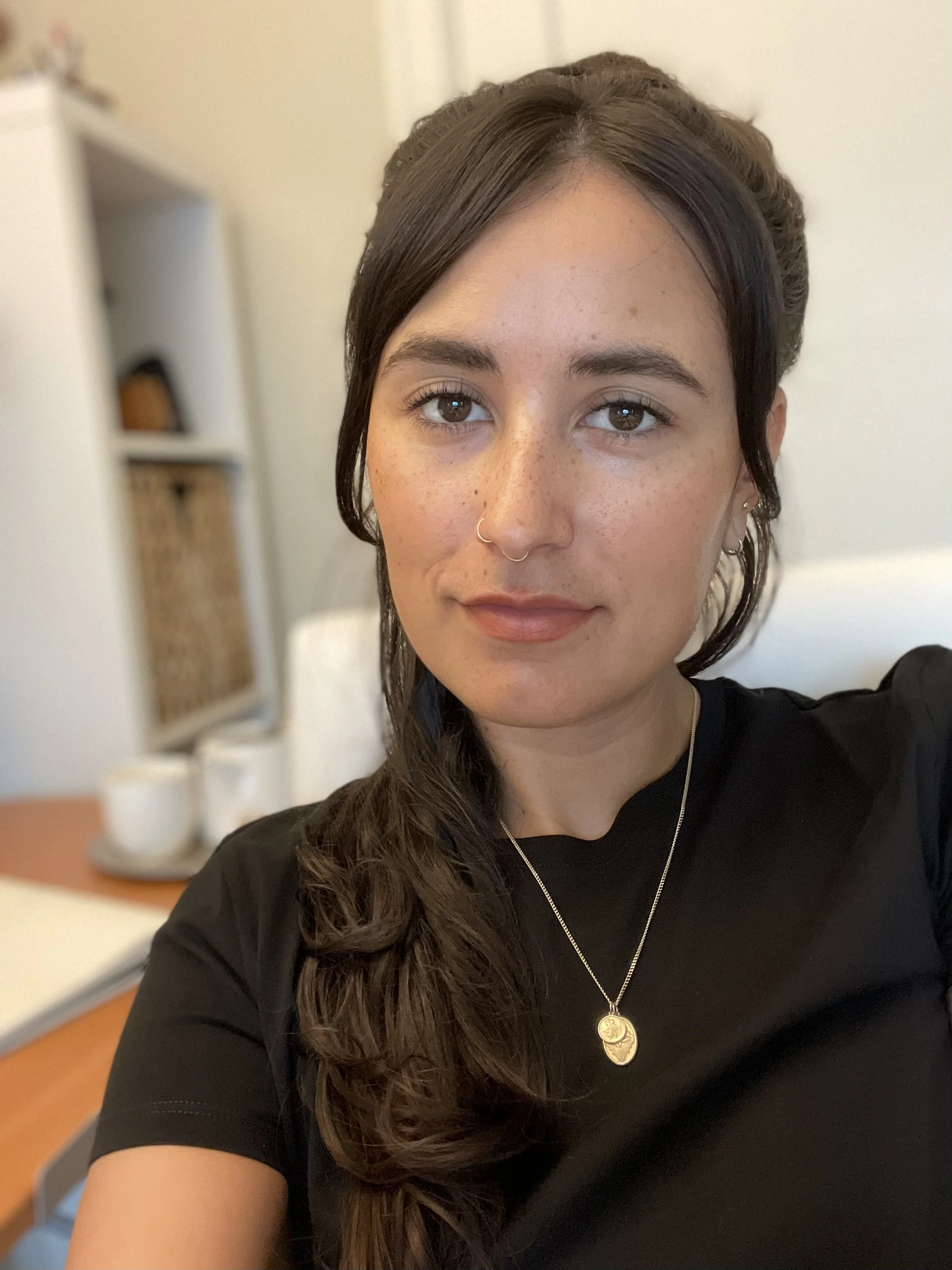 A young woman with long, wavy brown hair, wearing a black top, gold necklace, and nose ring, taking a selfie indoors.