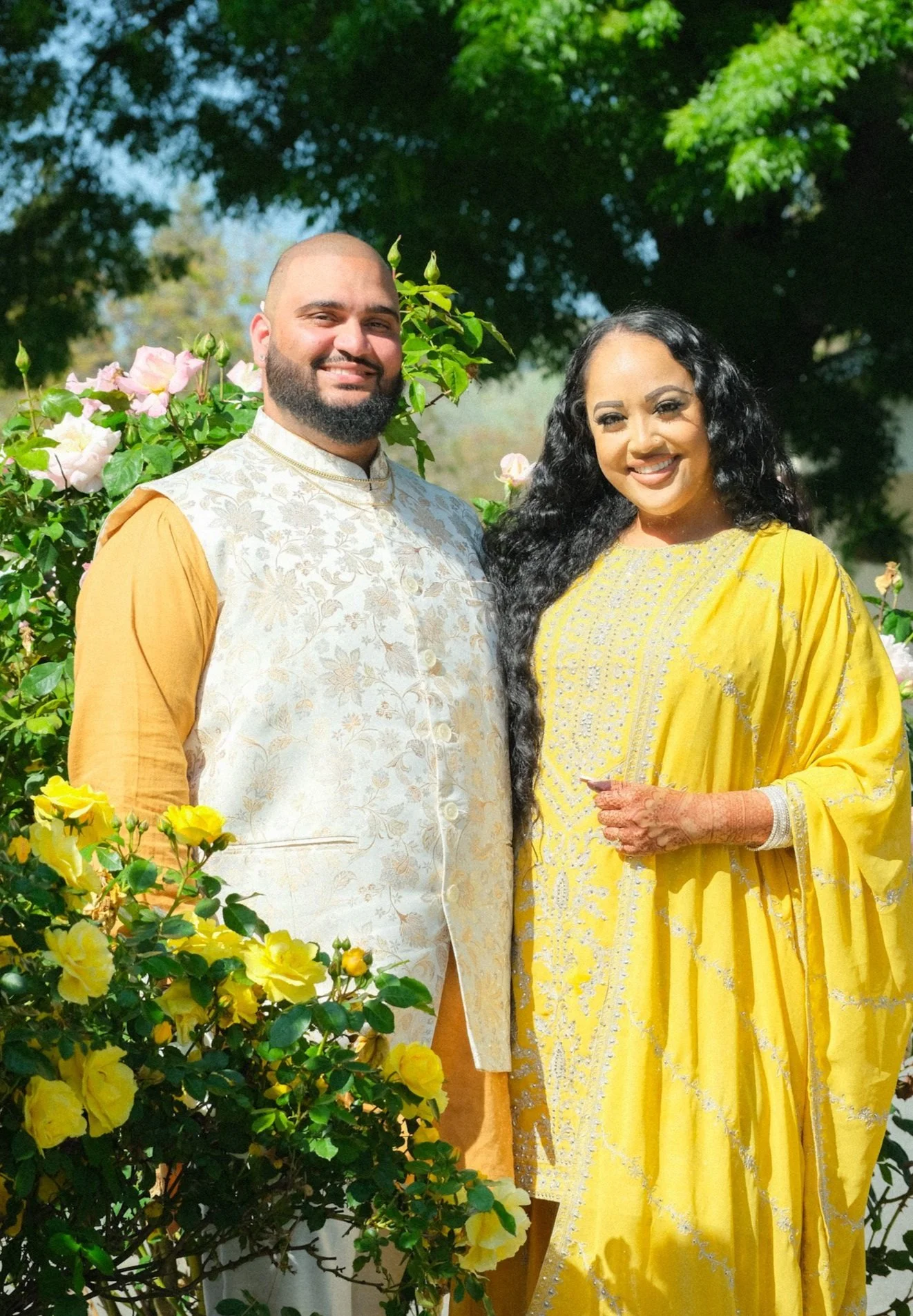 A smiling couple dressed in traditional South Asian attire, standing outdoors among yellow and pink flowers with green trees in the background.