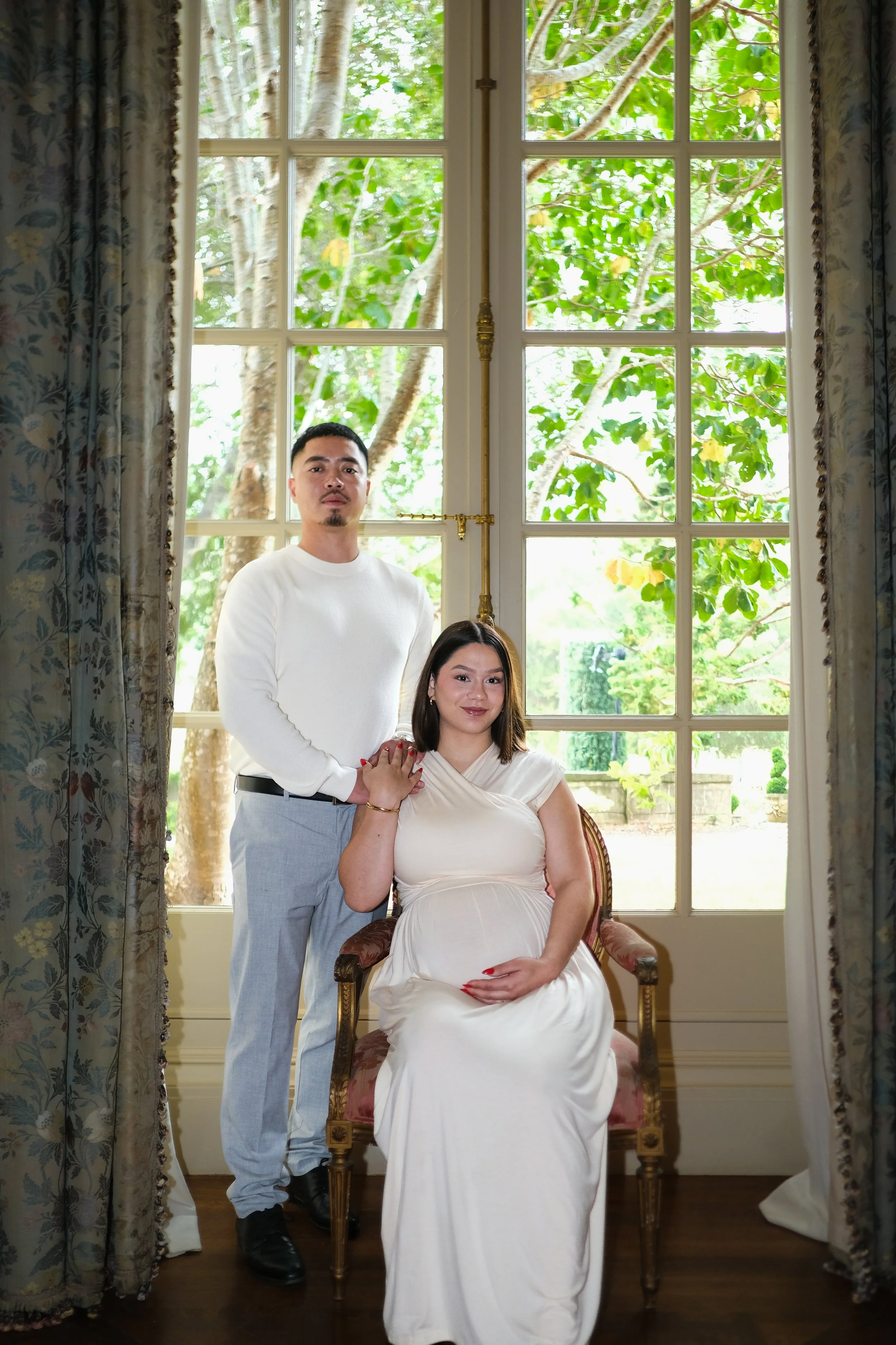 A pregnant woman sitting on a chair with her hand on her belly, and a man standing beside her with his hand on her shoulder, inside a room with large windows and floral curtains, overlooking a green garden.