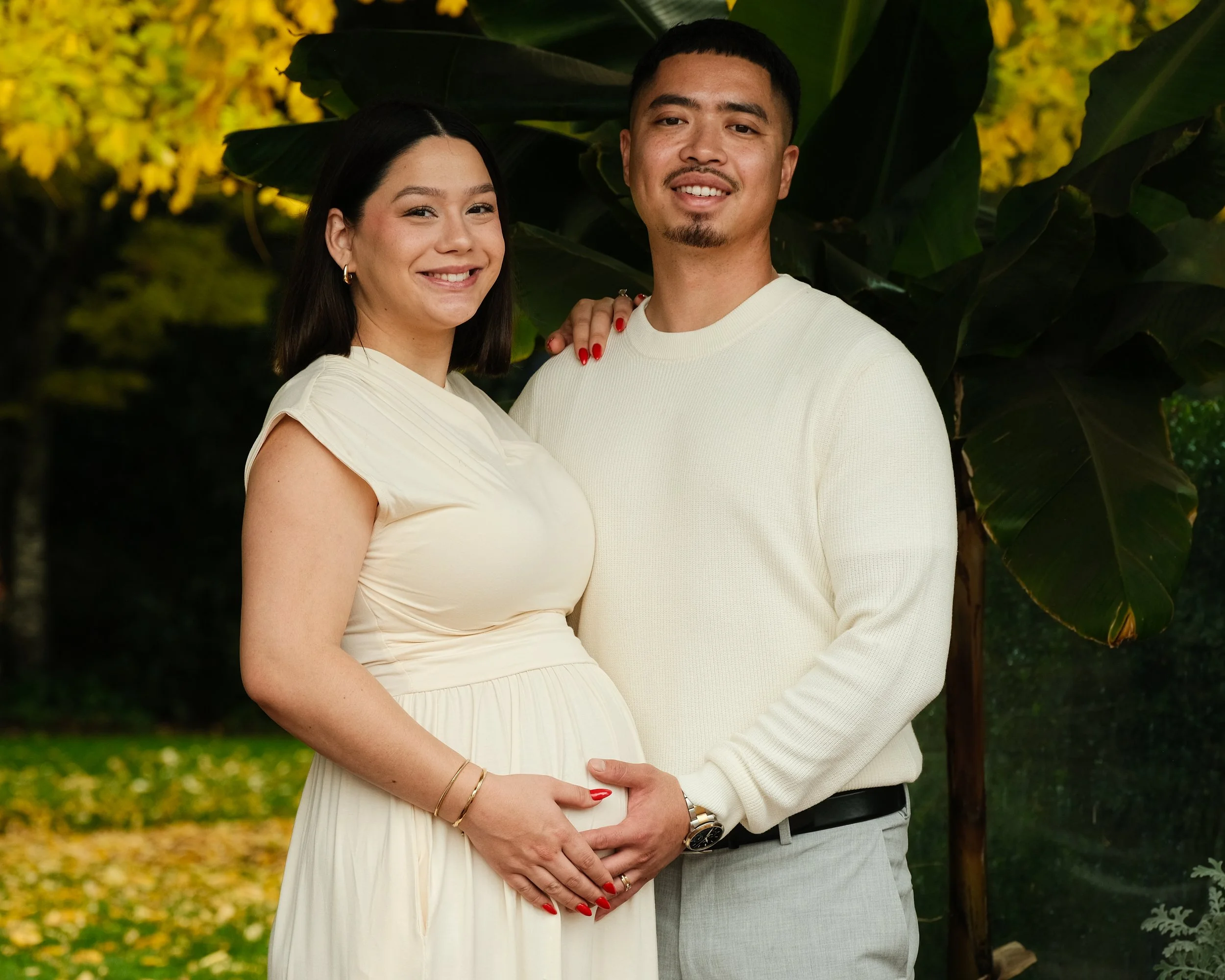 A smiling pregnant woman and a man stand outdoors among greenery, with the woman holding her belly as they pose for a photo.