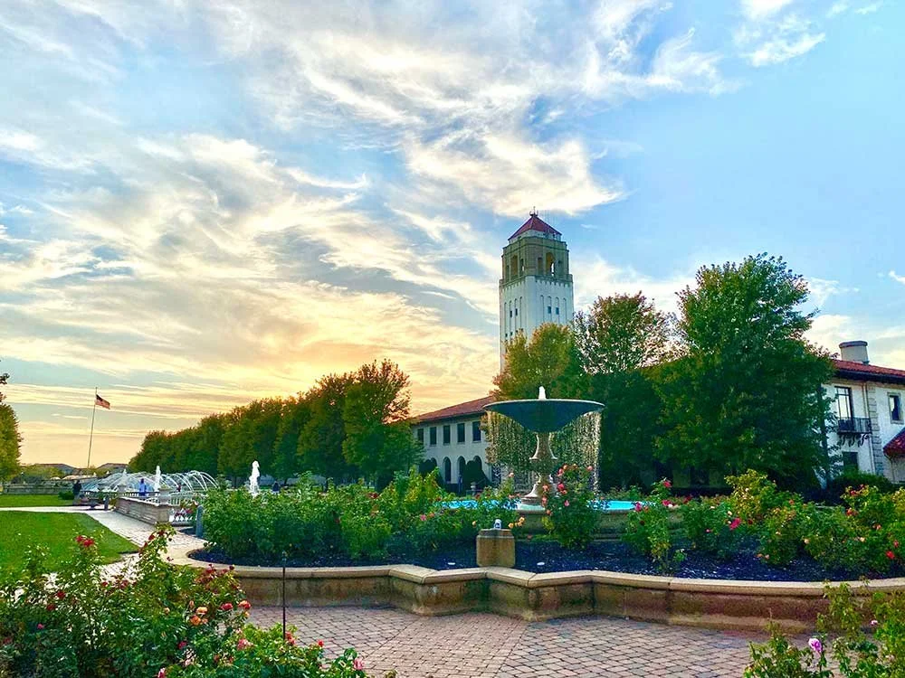 View of Unity Village's landscaped garden with a large central fountain, flowers, and trees. Buildings with red-tiled roofs and a tower are in the background. The sky is partly cloudy with a sunset glow.