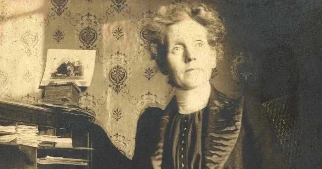 Vintage photo of Myrtle Fillmore in Edwardian attire standing beside a table with papers and a small framed picture, in a room with patterned wallpaper.