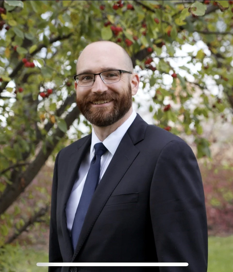 A smiling man with glasses, a beard, and a bald head, dressed in a dark suit and tie, standing outdoors in front of a tree with red berries and green leaves.