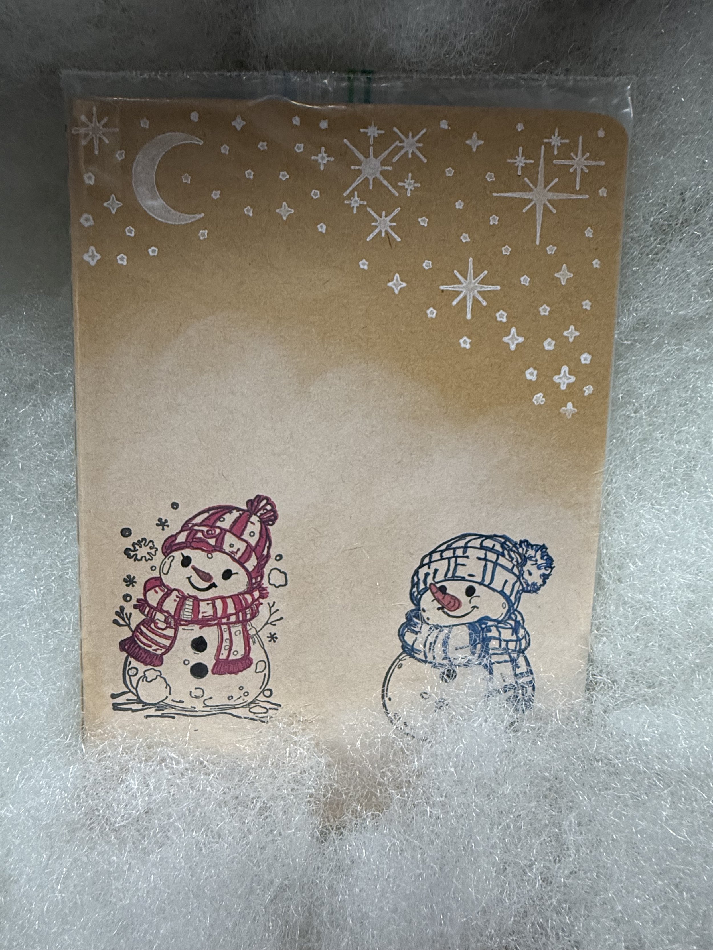 A small book nestled in fake snow. The cover features two round snowpeople in hand-colored hats and scarves. Above them is a starry winter sky.