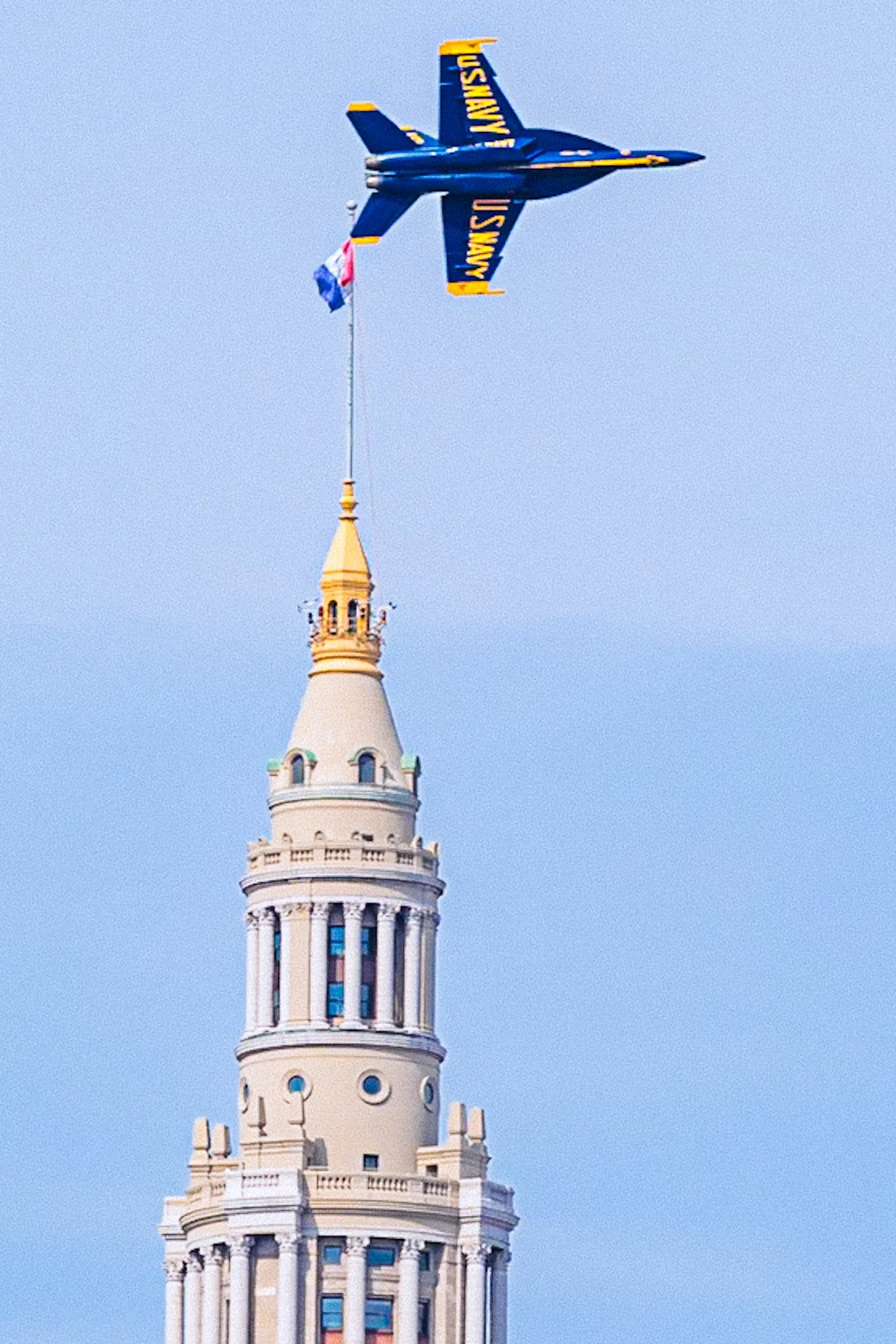 The top of a white tower with decorative features, topped with a golden cone, and a blue and yellow U.S. Navy jet flying above it, pulling a small American flag and a small French flag in the sky.