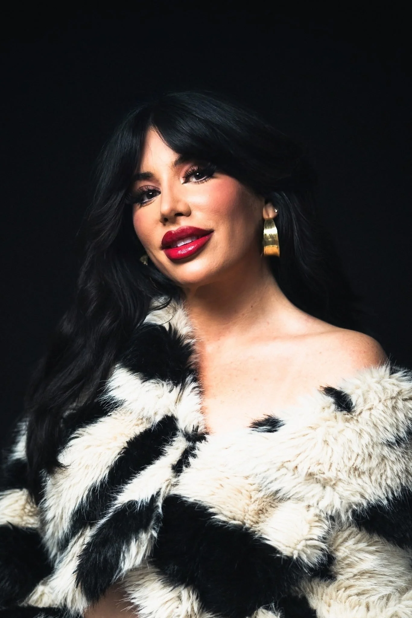 A close-up portrait of a glamorous woman with long, wavy black hair and bold bangs, featuring dramatic makeup including thick eyeliner, long lashes, and bright red lipstick. She gazes directly at the camera with a confident expression, wearing large 