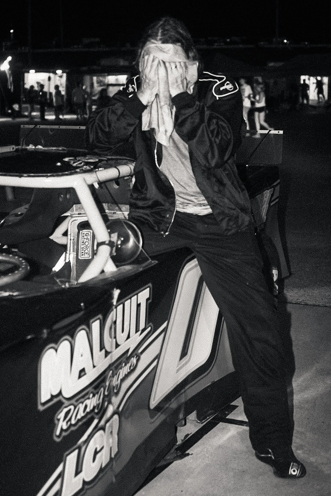 Person wearing headphones with hands covering face, standing near racing bike with logo on it at night.