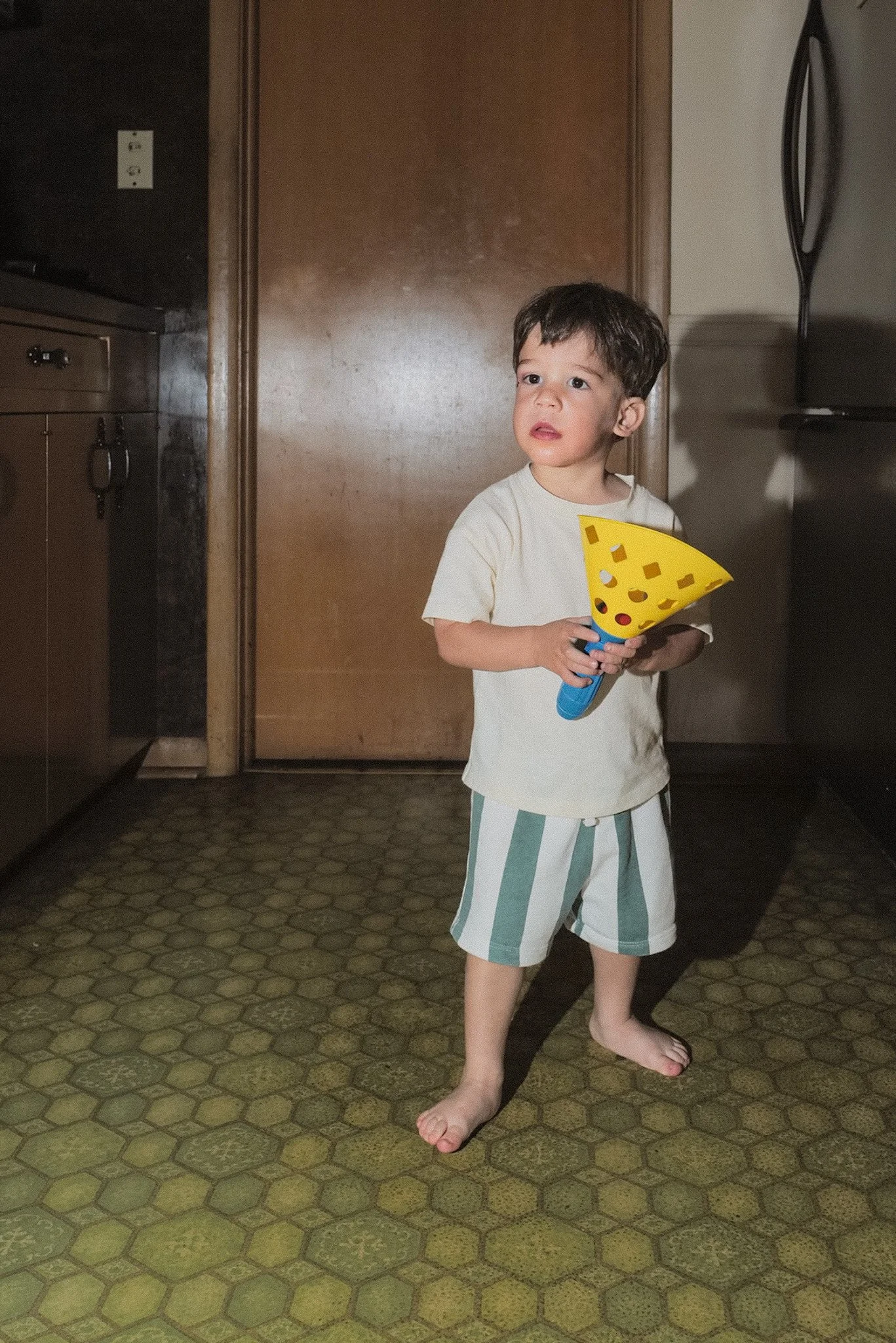 A young boy with dark hair, no shoes, wearing striped shorts and a t-shirt, stands barefoot on a patterned yellow-green carpet in a kitchen, holding a yellow plastic cone with holes, looking off to the side.