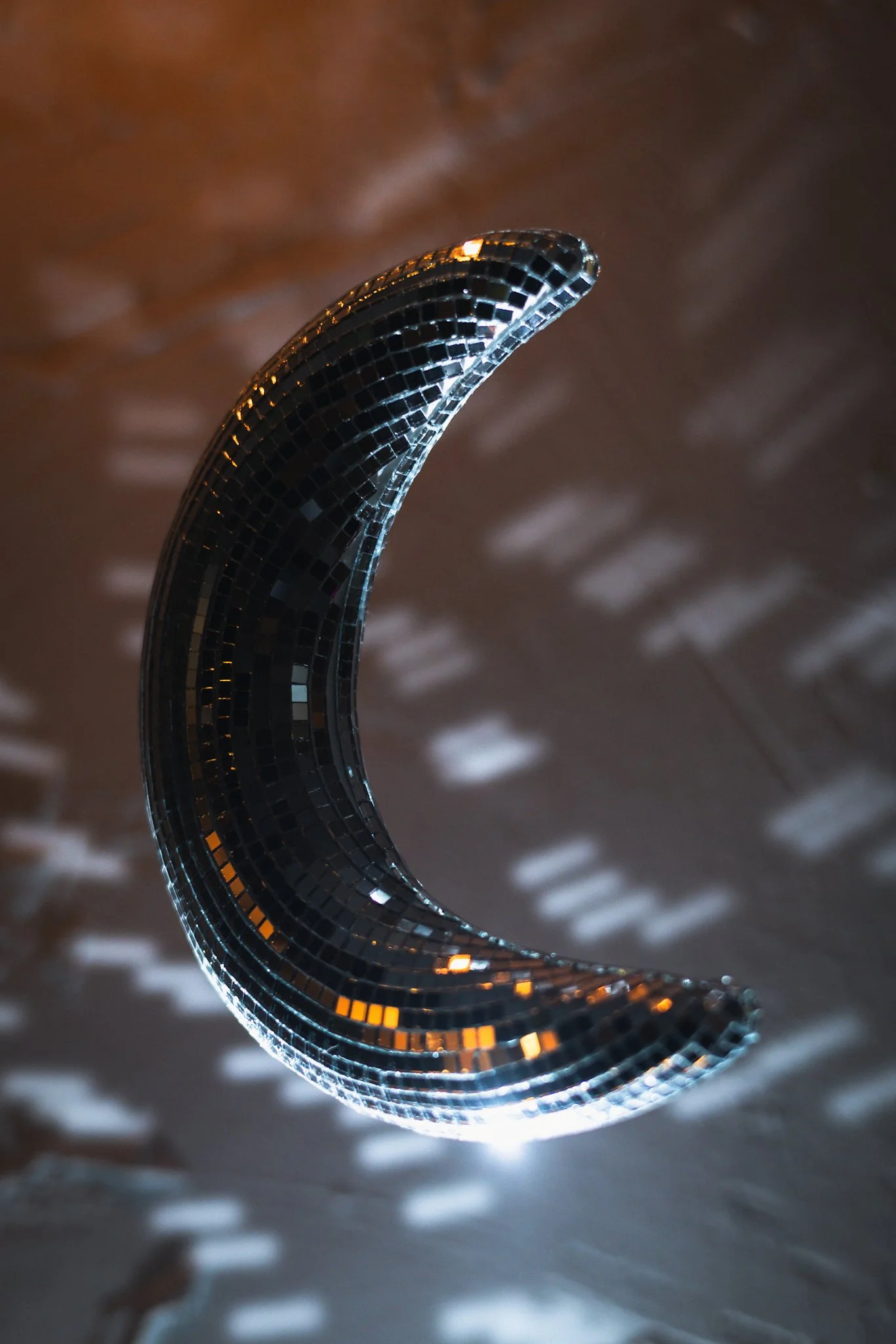A crescent moon–shaped disco ball made of small mirrored tiles hangs from a chain, reflecting soft white and warm orange light patterns onto a dark, out-of-focus background.