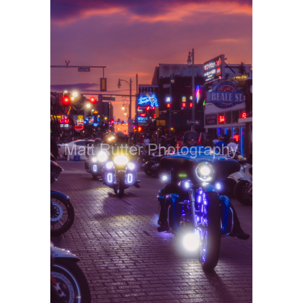 BIkers riding down Beale Street with lights on and a cotton candy sky in the background