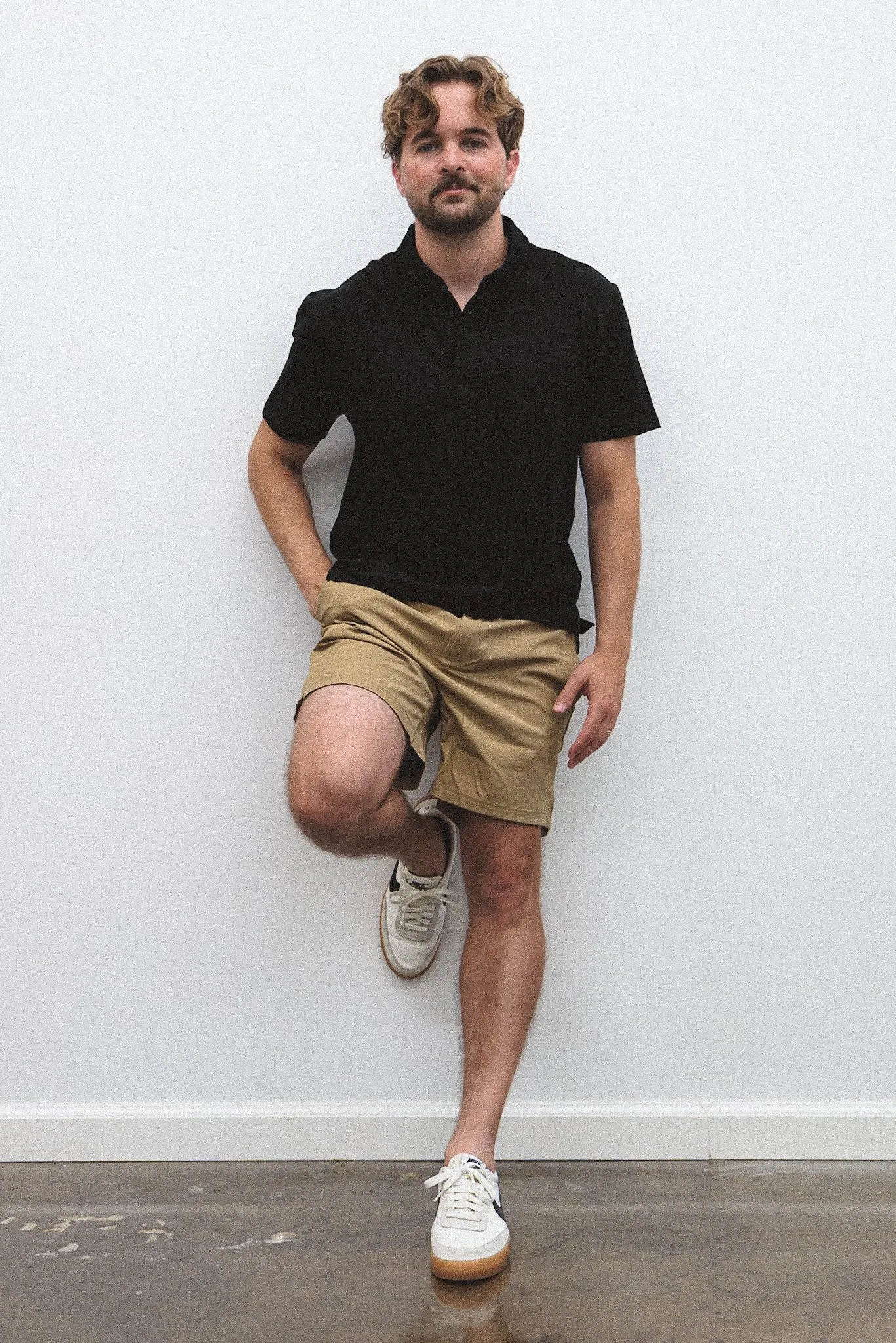 Young man with curly hair and beard wearing a black polo shirt, beige shorts, and white sneakers, leaning against a white wall with one foot raised and hand in pocket.
