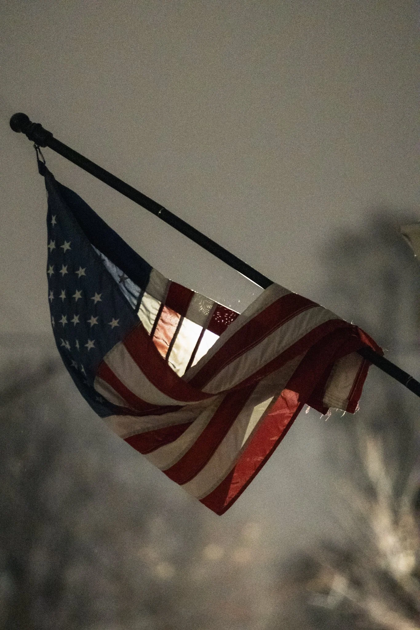 A weather-beaten American flag droops on a pole at night, its fabric stiff with ice, dimly lit against a soft, gray winter sky.