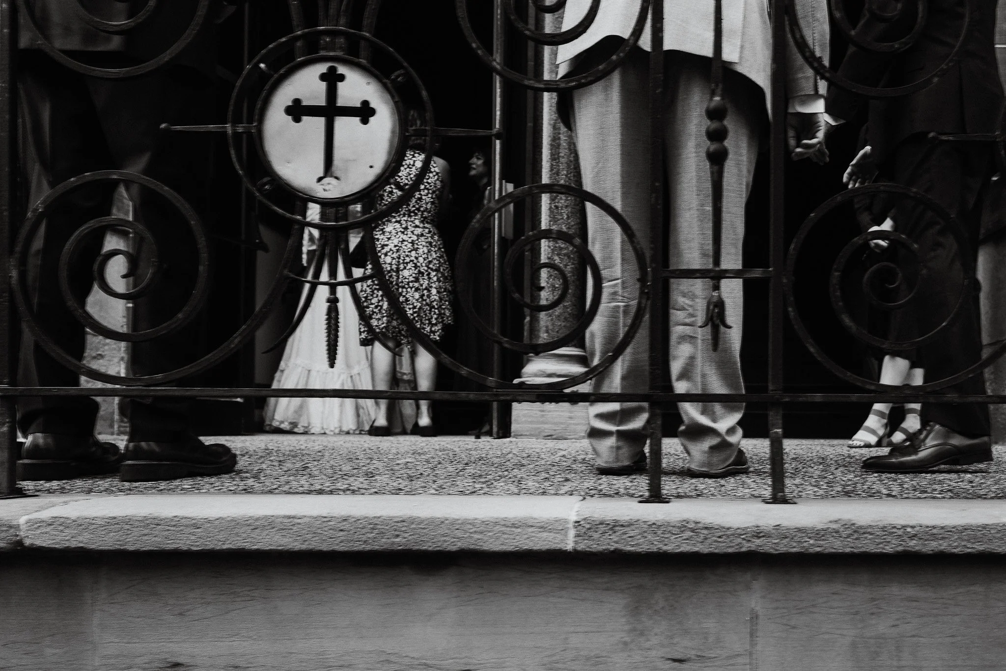 A black and white photo taken through a decorative wrought iron fence showing part of a church exterior with a clock and cross on the facade. People are walking by, with only their lower bodies visible.