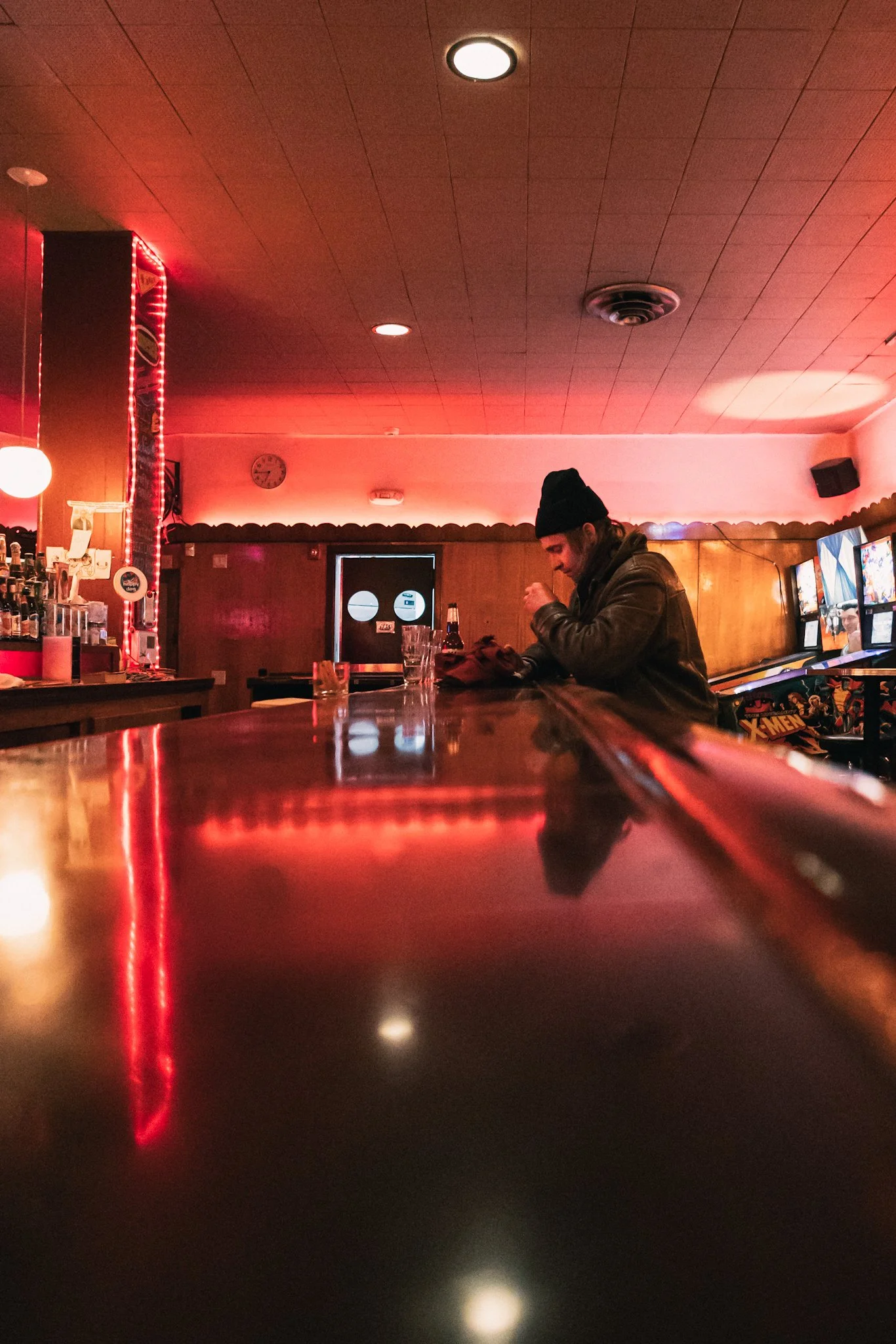 Low-angle view inside a dimly lit bar with red neon glow and warm lighting: a man in a beanie and jacket sits pensively at the polished wooden bar, gazing ahead amid reflections, glasses, barstools, and subtle background details like a clock and pool